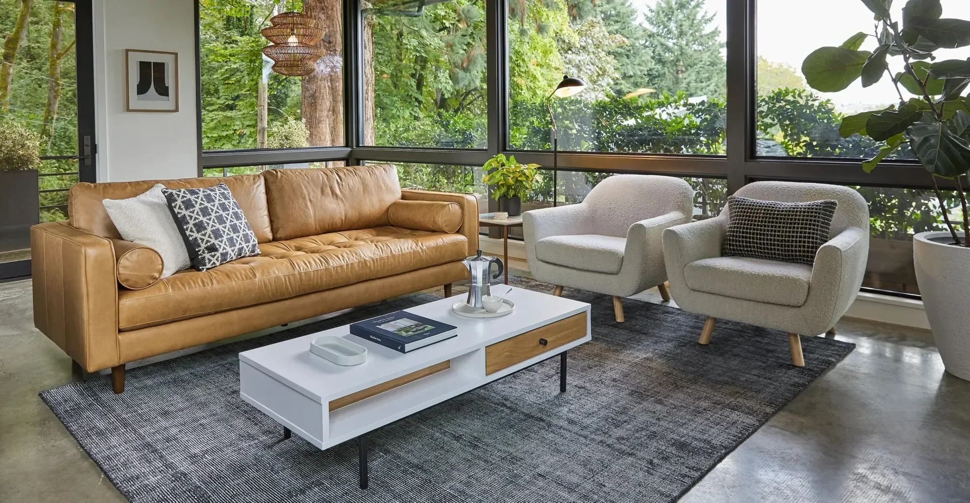 Living room with large windows, tan leather sofa, two light-colored armchairs with cushions, a modern white coffee table with wooden accents, a small side table with a lamp and potted plant, and green foliage outside the window.