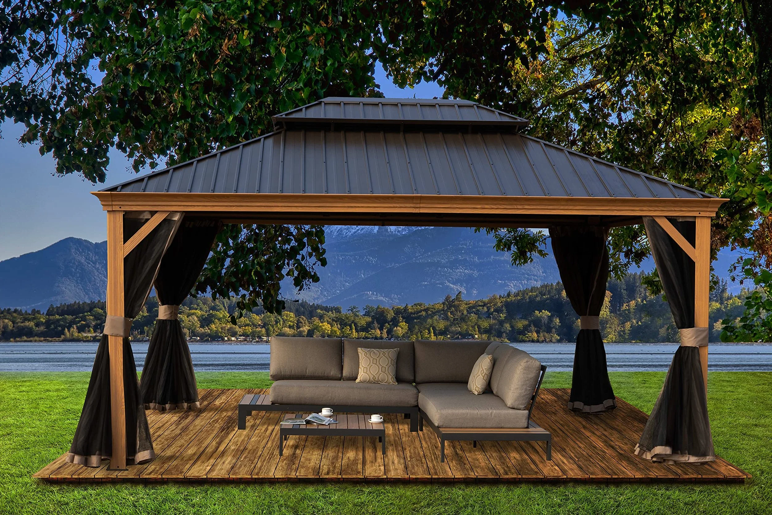 Outdoor gazebo with a wooden deck, black curtains, and seating area, overlooking a lake with mountains in the background.