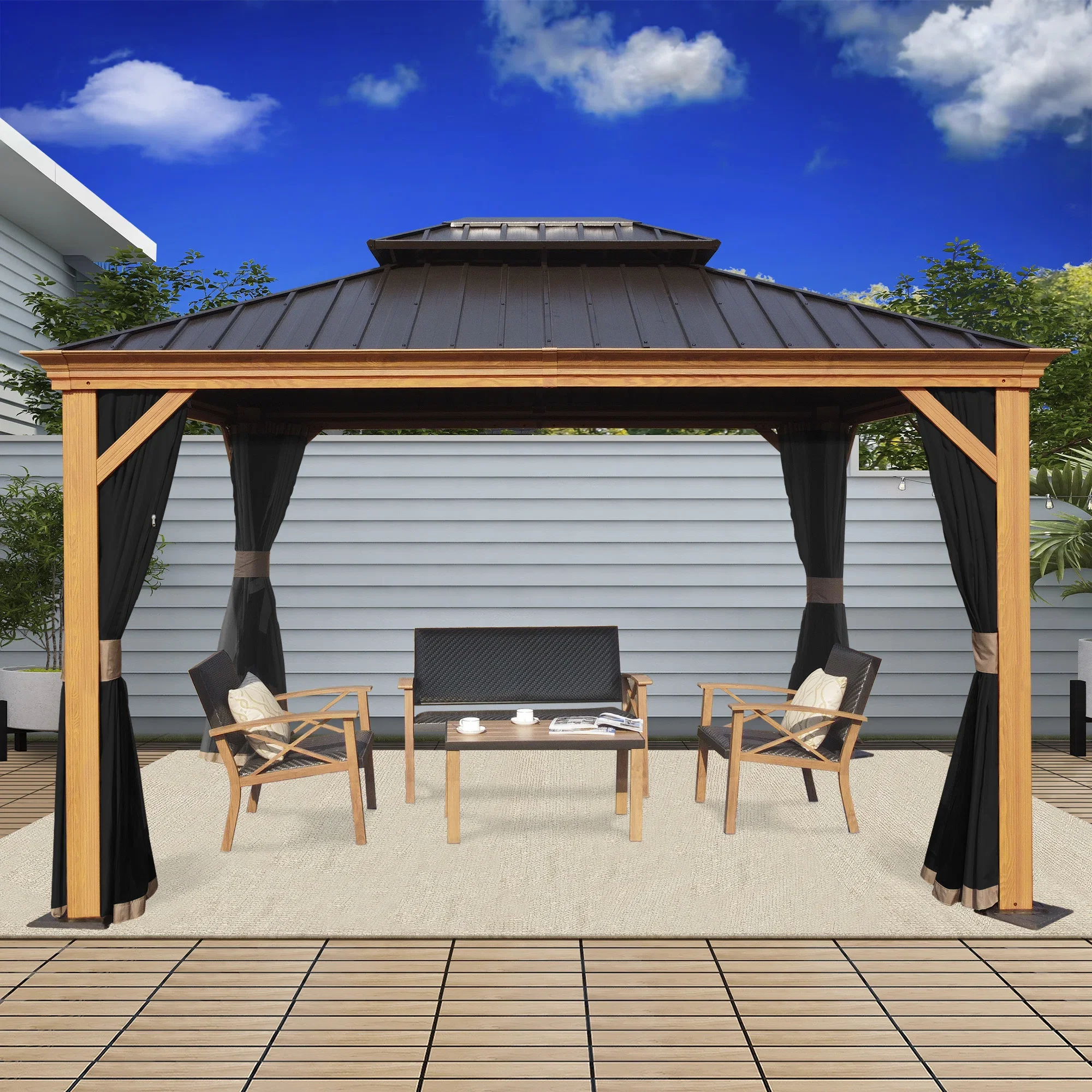 A backyard patio featuring a wooden gazebo with black curtains, patio furniture with four chairs around a small table, a rug underneath, and a background of a house with siding and greenery, under a blue sky with clouds.