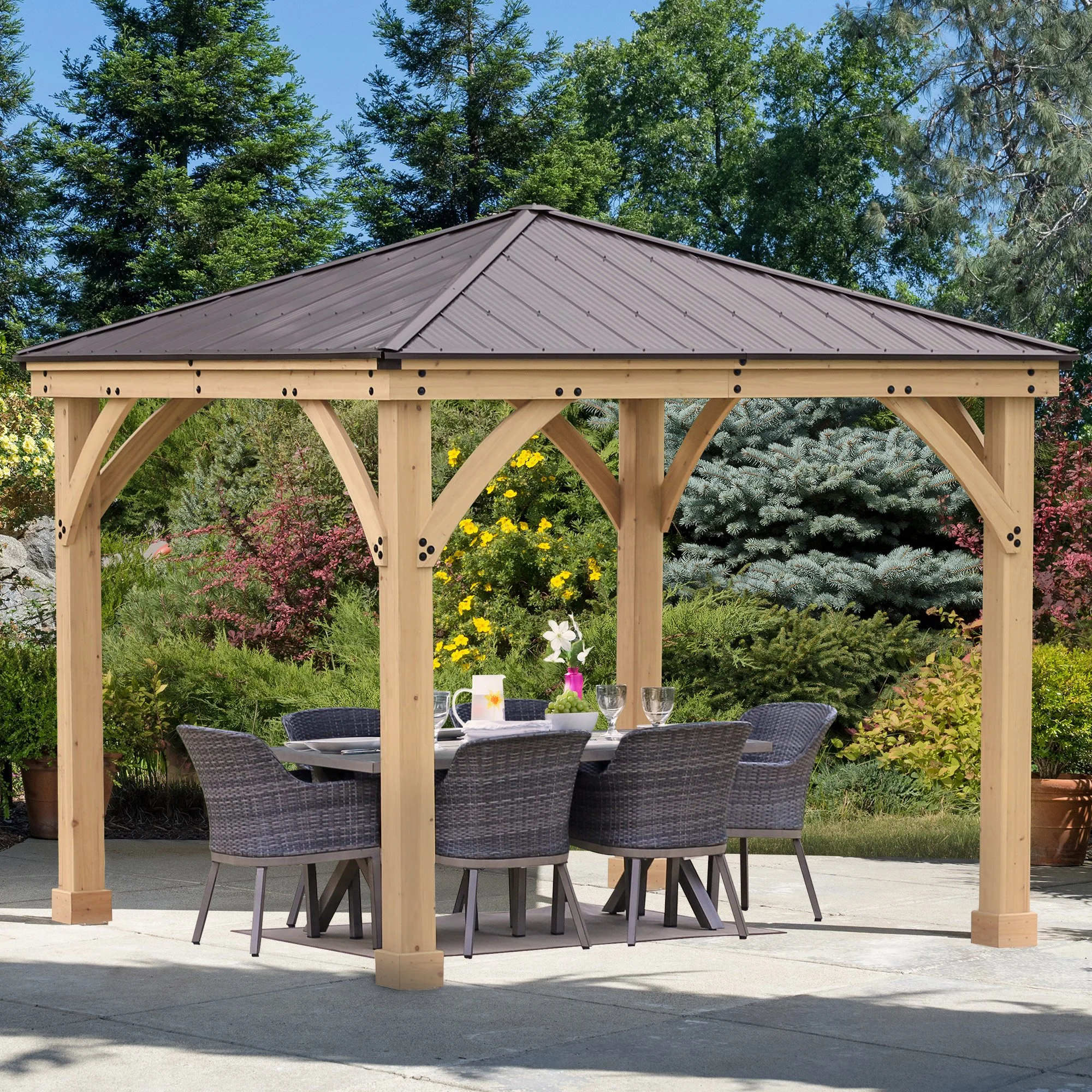 Wooden outdoor gazebo with a metal roof, surrounding a dining table with six chairs, set with glassware and a flower centerpiece, in a garden with trees and colorful bushes.