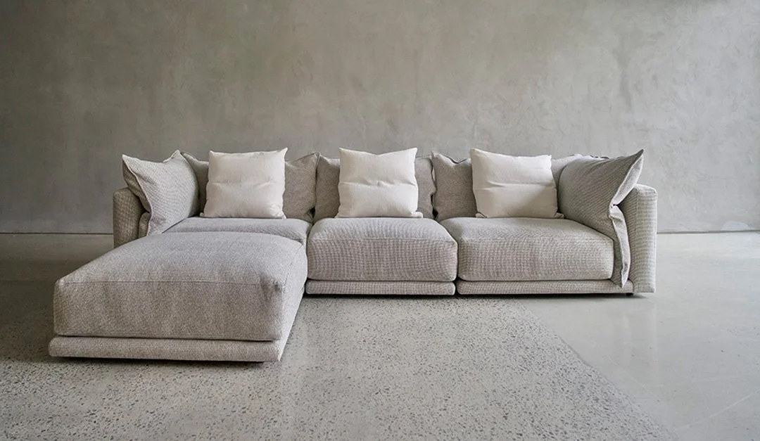 A light gray sectional sofa with tiered back cushions and a chaise at one end, placed against a textured light gray wall on a neutral speckled floor.
