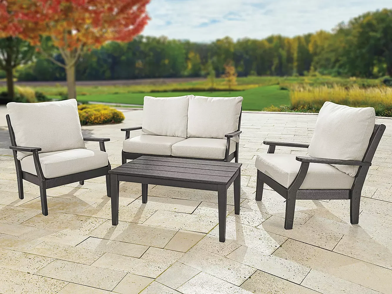 Outdoor patio furniture set including two armchairs, a two-seater loveseat, and a small rectangular table, with a green park and trees in the background.