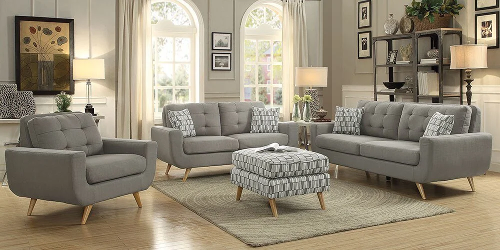 Living room with gray sofas, patterned cushions, a matching ottoman, a beige area rug, and shelves with decorative items, vases, and lamps.