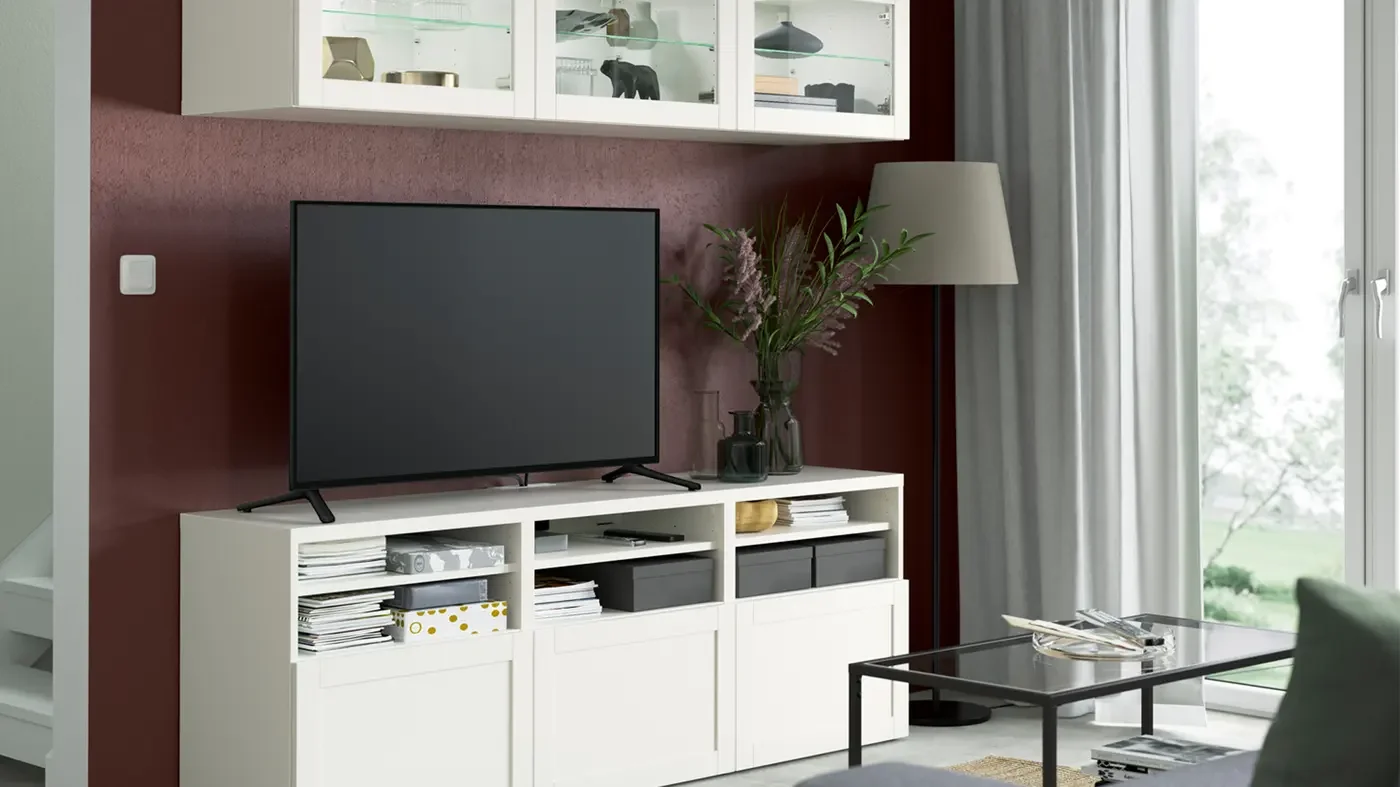 Living room with a white TV stand, a flat-screen TV, a tall floor lamp, a vase with flowers, coffee table, and sliding glass door with curtains.