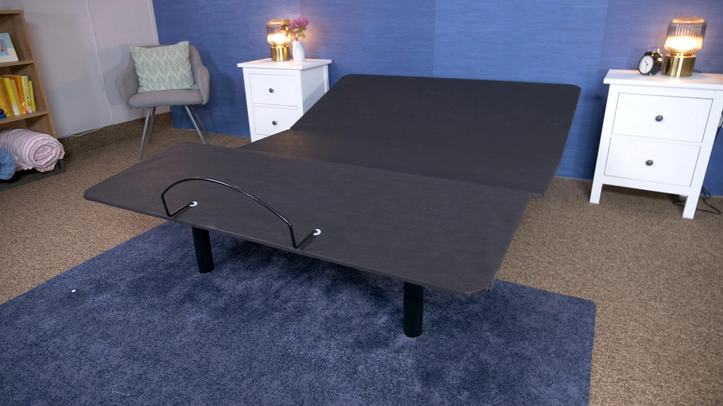 A dark wooden massage or therapy table with a curved handle on one end, placed in a room with beige carpet and two white nightstands with lamps and a clock. A gray upholstered chair with a cushion and a bookshelf with books and blankets are also visible.