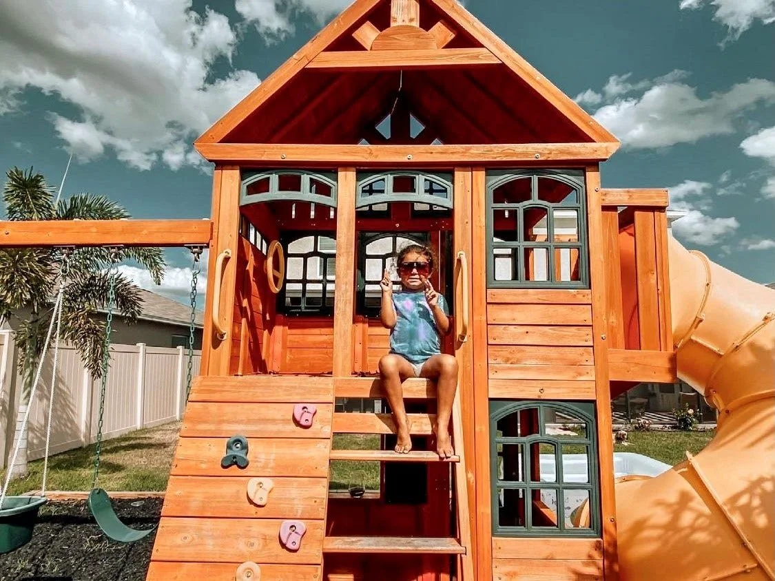 Child sitting on a porch of a wooden playhouse with a slide, wearing sunglasses, with a swing set to the left and a white fence in the background.