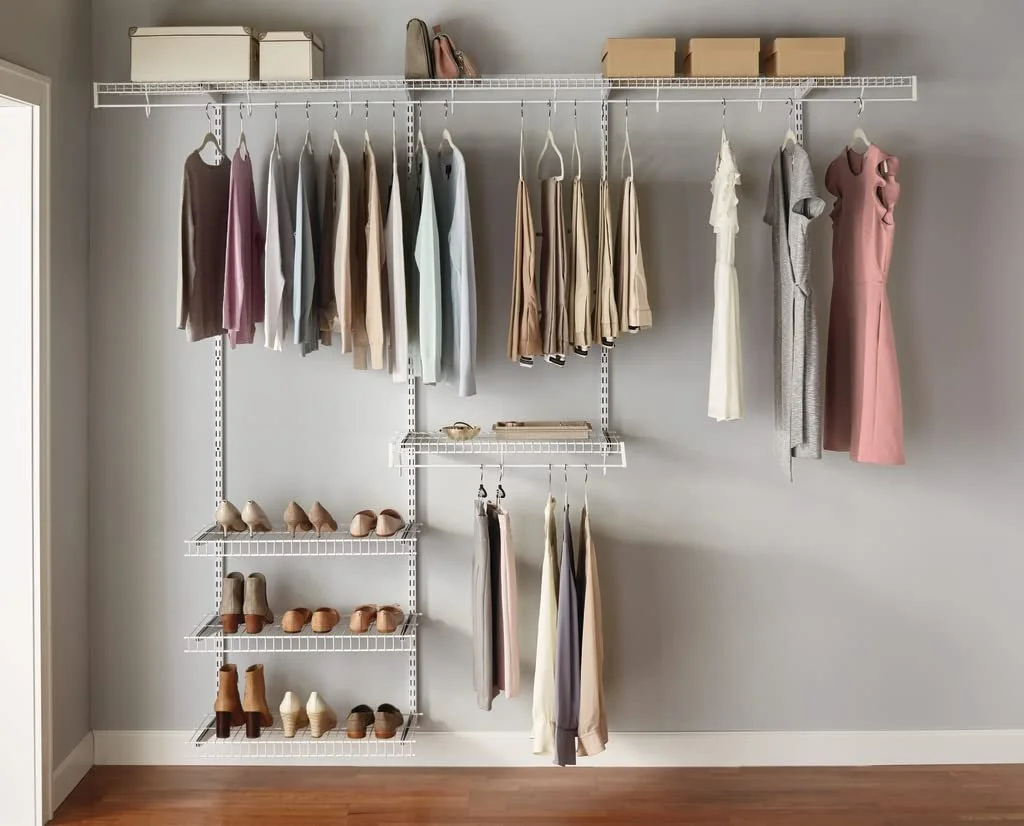 A neatly organized closet with white wire shelving holding various folded clothes, shoes, and storage boxes against a gray wall and wooden floor.