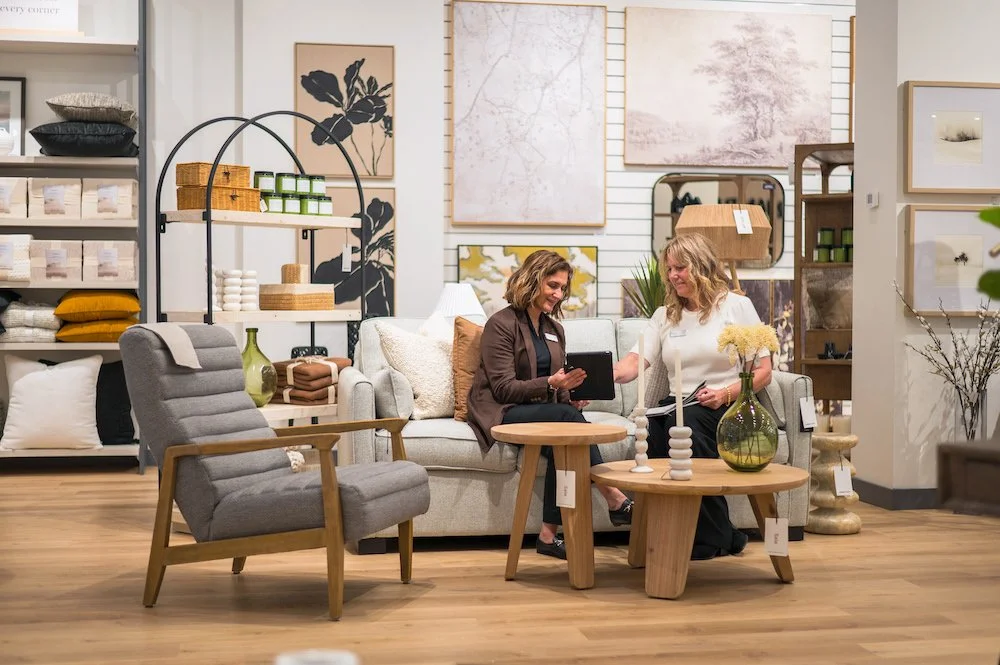 Two women shopping for home decor in a furniture store. They are sitting on a light-colored sofa, looking at a tablet. The store has shelves with cushions and folded fabrics, and walls decorated with framed artwork and mirrors. A gray armchair and a round wooden coffee table with decorative items, including candles and vases, are in the foreground.