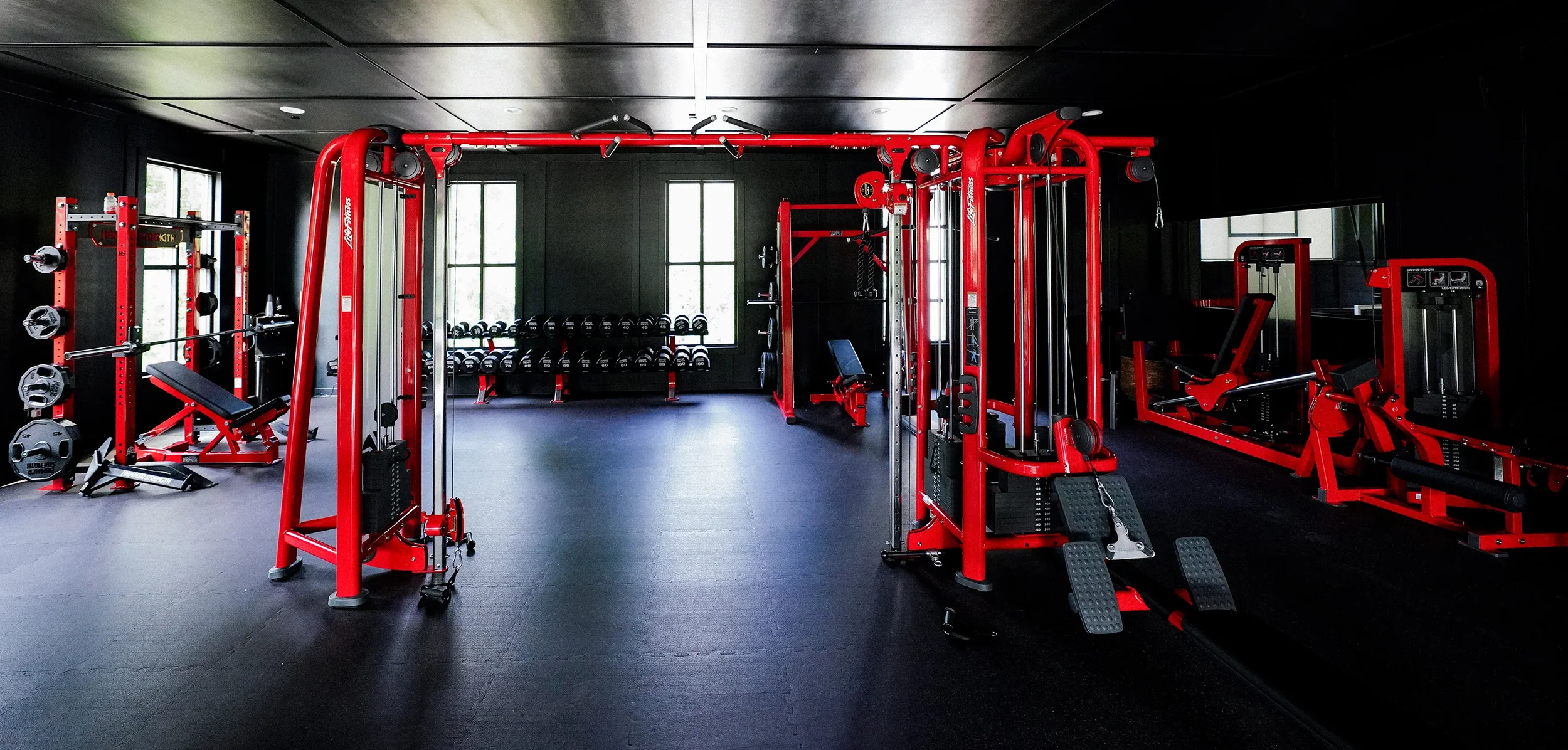 Interior of a modern gym with black walls and floors, featuring red exercise equipment including weight machines, a bench press, and free weights, with windows letting in natural light.