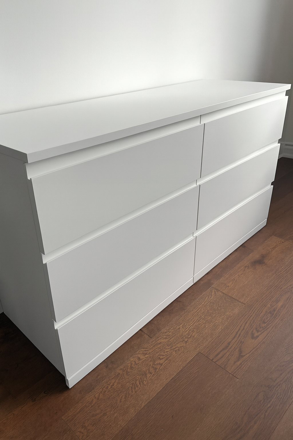 A white modern dresser with six drawers in a room with wooden flooring.