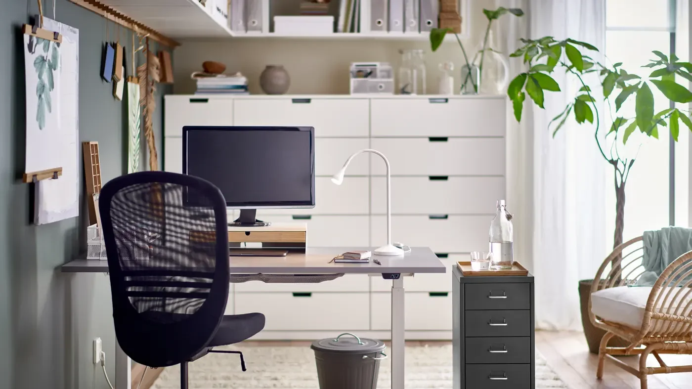 Home office with a desk, computer monitor, black office chair, white desk lamp, and supply cabinet, with plants and natural light.