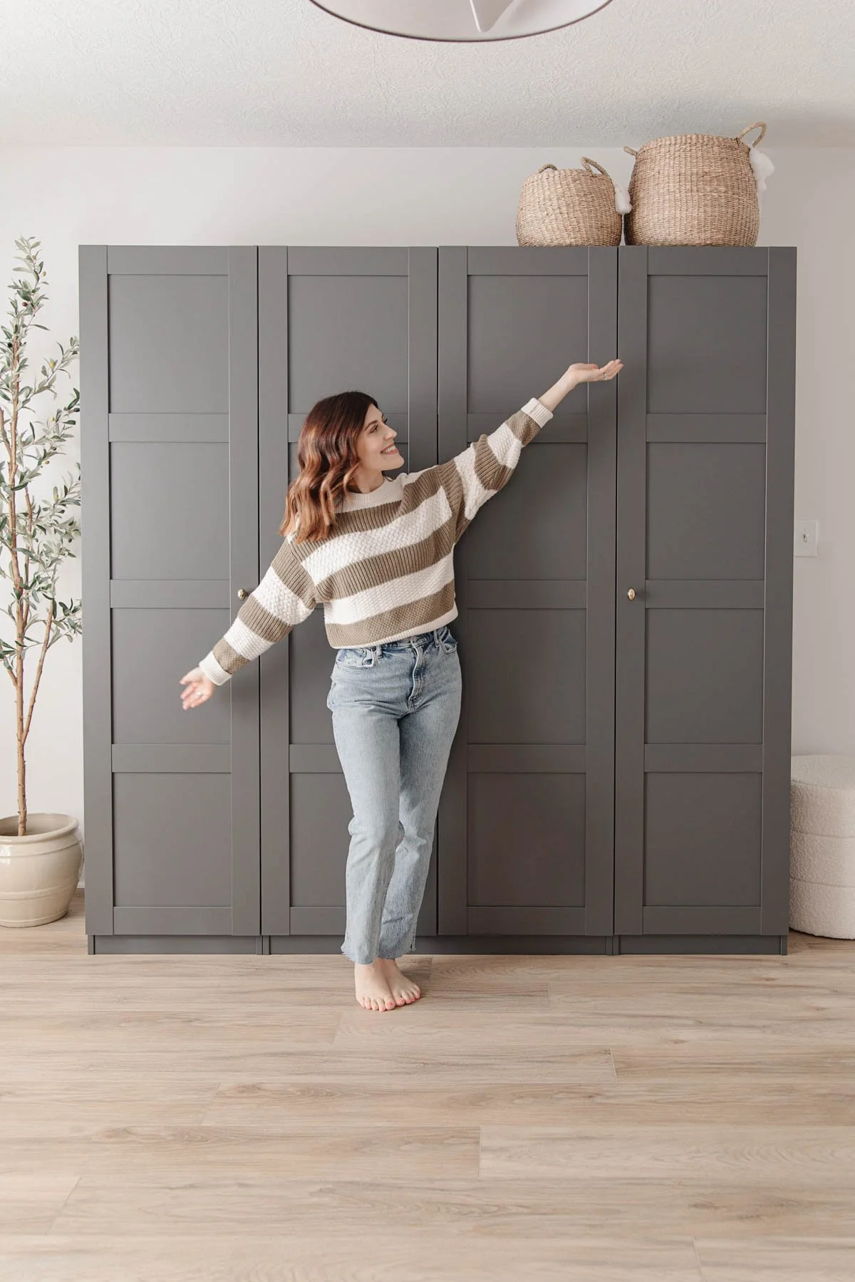 A woman standing barefoot on a light wood floor, reaching up towards a large dark gray cabinet with woven baskets on top, inside a bright room.