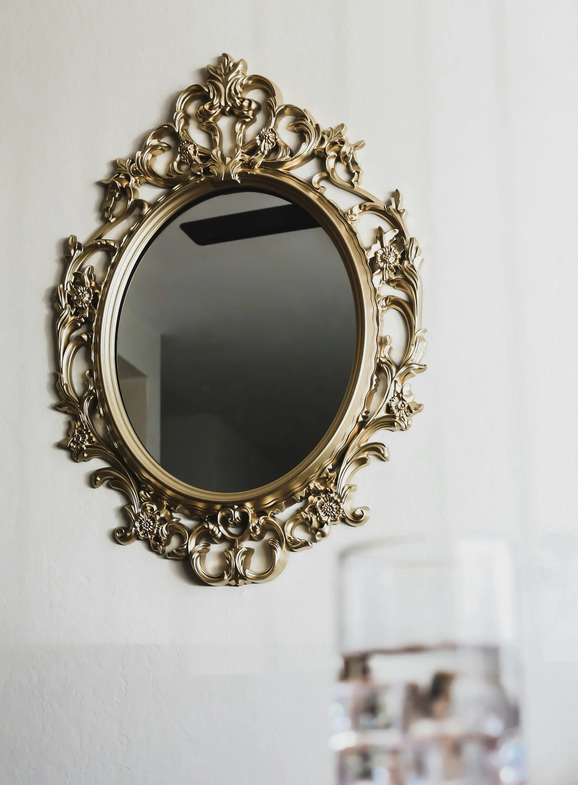 A decorative gold-framed oval mirror hanging on a white wall, with a blurred glass object in the foreground.