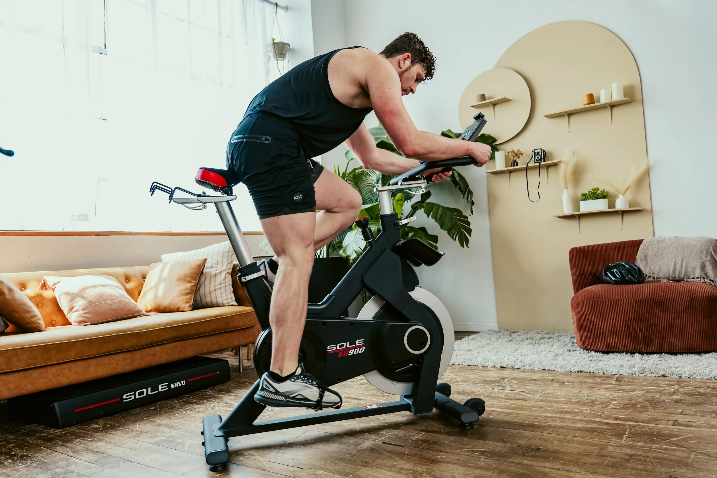 A man exercising on a stationary bike indoors, wearing black athletic shorts and a tank top, in a living room with a sofa, plants, and decorative shelves.