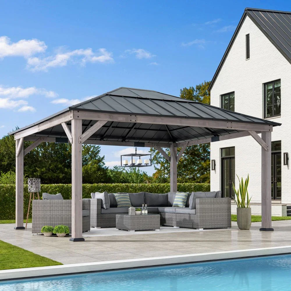 Outdoor patio with a wooden gazebo, outdoor seating with gray wicker furniture, a pool, and a white house with black window frames under a blue sky.