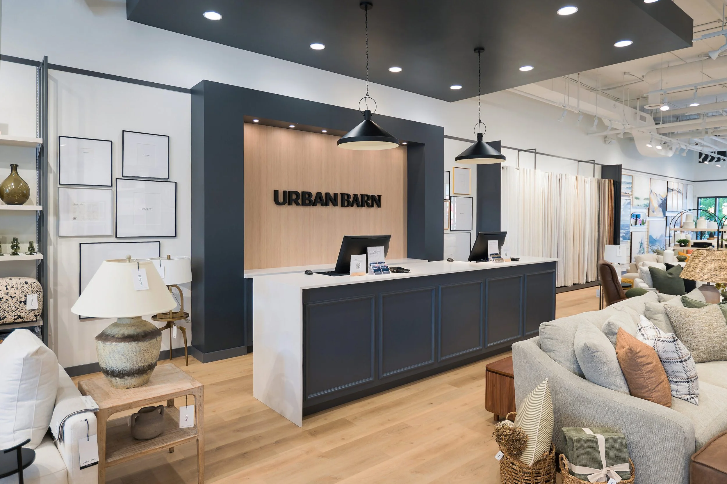 Modern furniture store interior with a dark blue and wood-paneled reception desk displaying the sign "Urban Barn," surrounded by sofas, lamps, and home decor items.