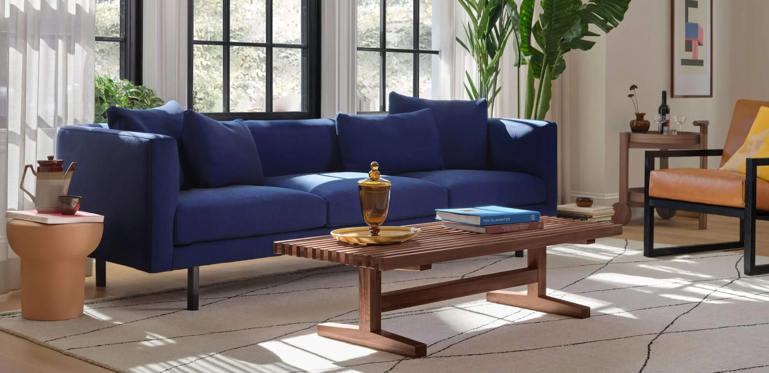 Living room with a blue sofa, a wooden coffee table holding glassware and books, a kidney-shaped side table with a teapot and cup, a tan and black armchair, and a large green plant near windows with white curtains, sunlight casting shadows on a patterned rug.