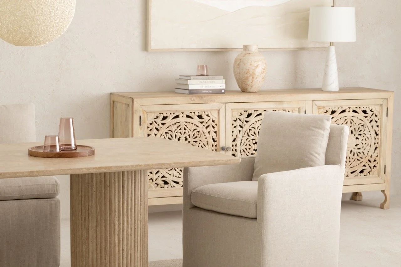 A minimalist, neutral-colored living room corner featuring a wooden table with two pink glass cups on a round wooden tray, a cream armchair, and a light wood sideboard with intricate carved door panels. On the sideboard are a stack of books, a large vase, a pink glass, and a white table lamp. Wall art and textured walls complement the decor.