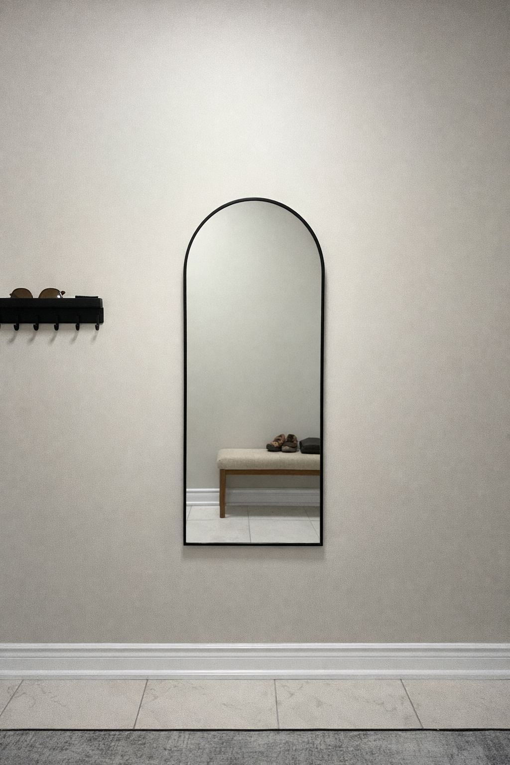 A tall, arched mirror on a plain beige wall, reflecting a small beige bench with two pairs of shoes and a black case on top.