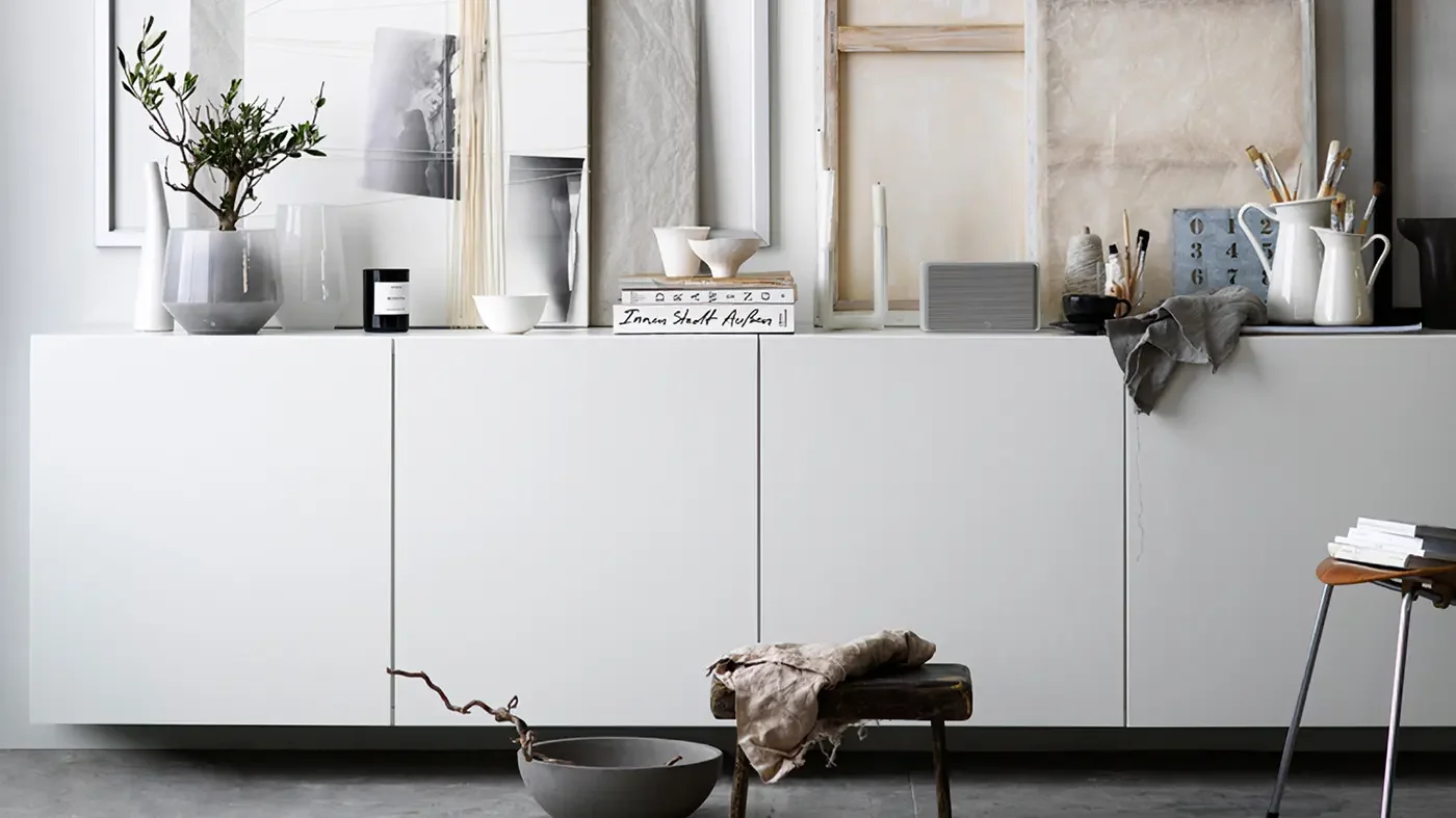 A minimalist white kitchen counter with decorative items, framed artwork, and art supplies, along with a small rustic stool with a cloth on it and a black bowl on the floor.