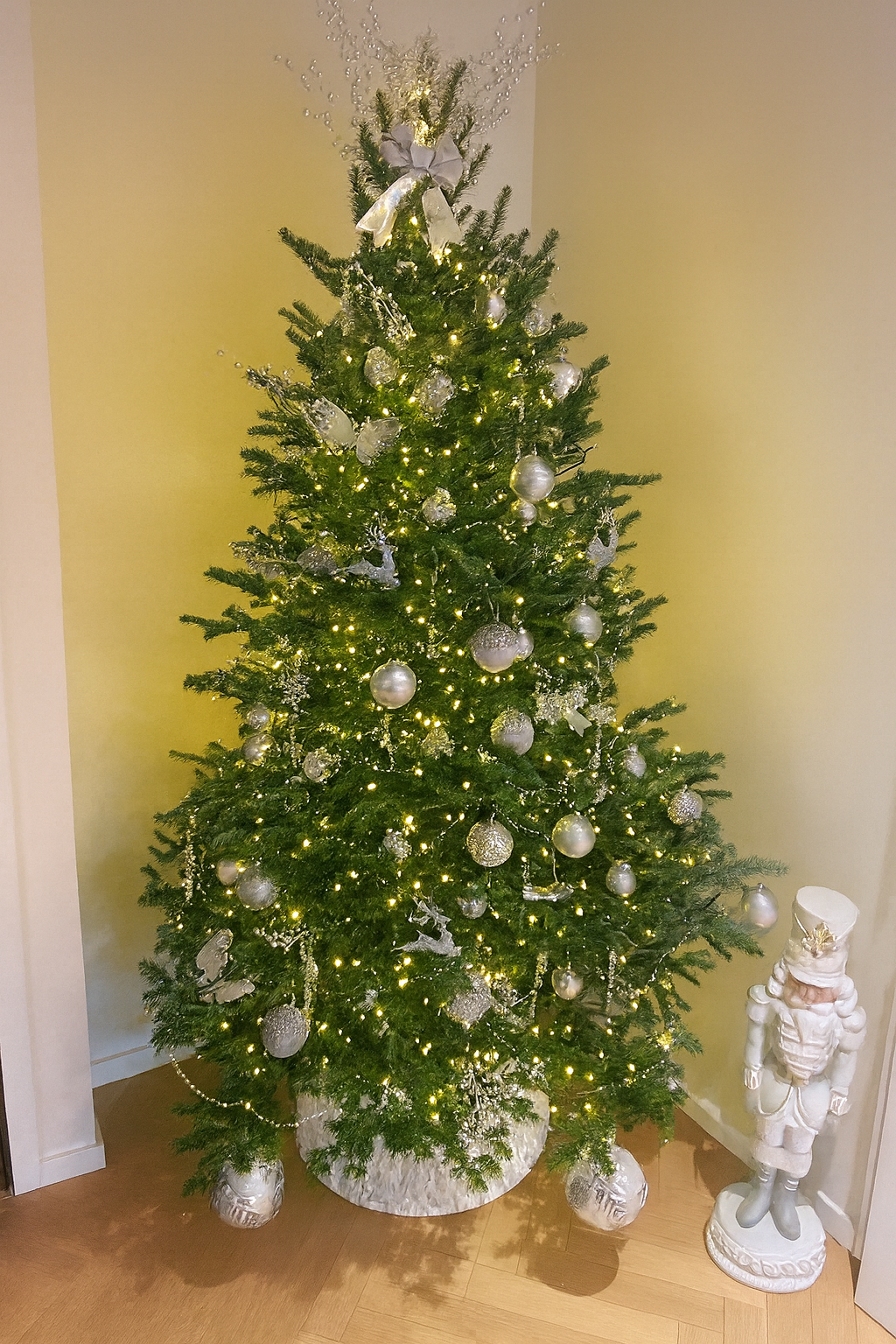 Christmas tree decorated with white and silver ornaments, lights, and a large bow at the top, with a nutcracker figure standing beside it.