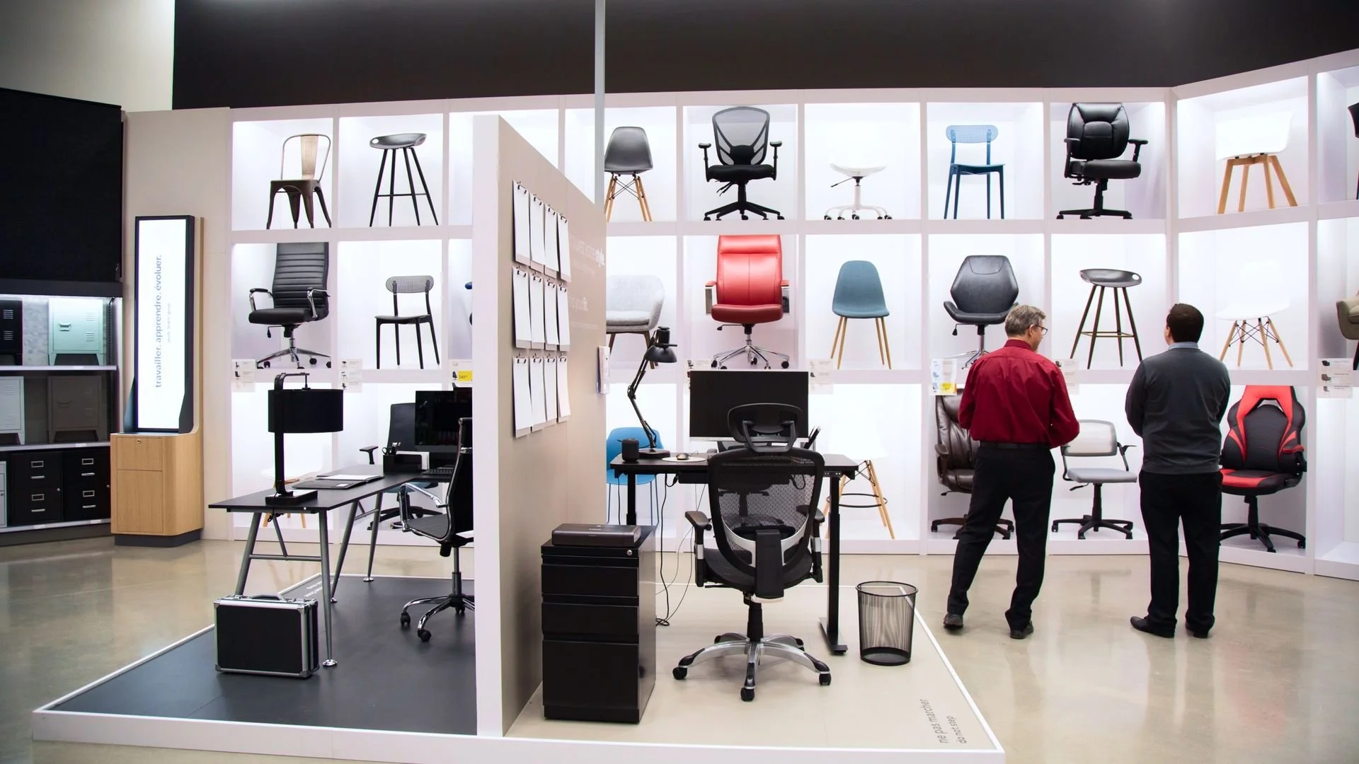 Showroom with various office chairs on shelves and demo stations, two men browsing and talking, modern office furniture display.