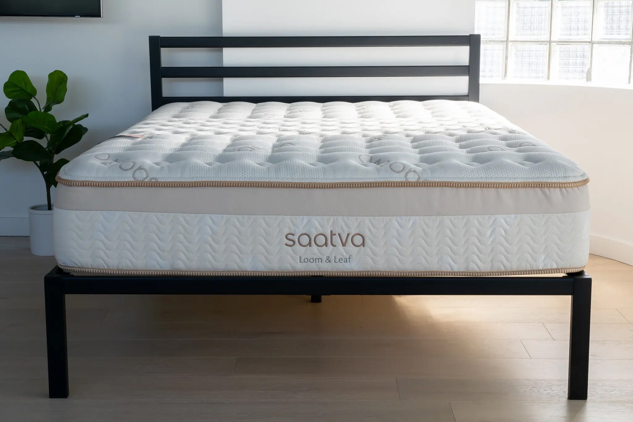 A bed with a black metal frame and a mattress labeled 'Saatva Loom & Leaf' in a bedroom with a large window and a potted plant in the background.