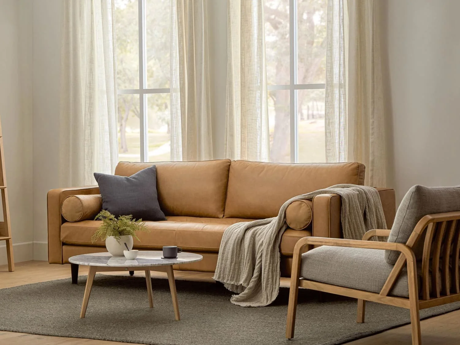 Living room with a tan leather sofa, gray armchair, round marble coffee table, and large windows with sheer curtains.