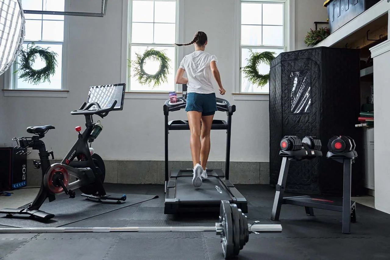 A woman running on a treadmill in a home gym, surrounded by workout equipment including a stationary bike, dumbbells, and a barbell on the floor, with large windows and holiday wreaths.