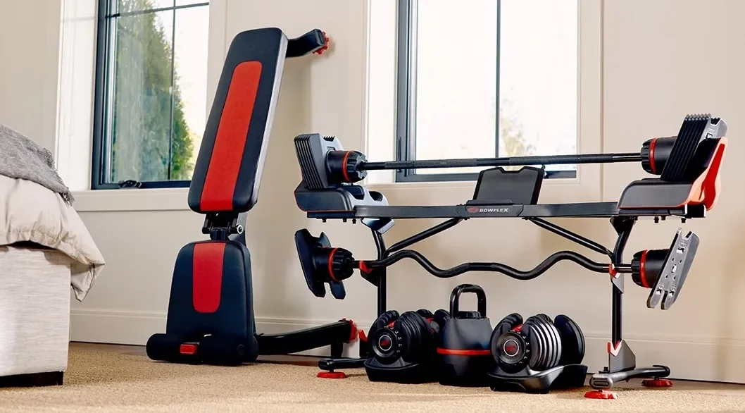 Indoor home gym equipment including a Bowflex adjustable bench, dumbbells, a kettlebell, and a weight plate storage rack near window.