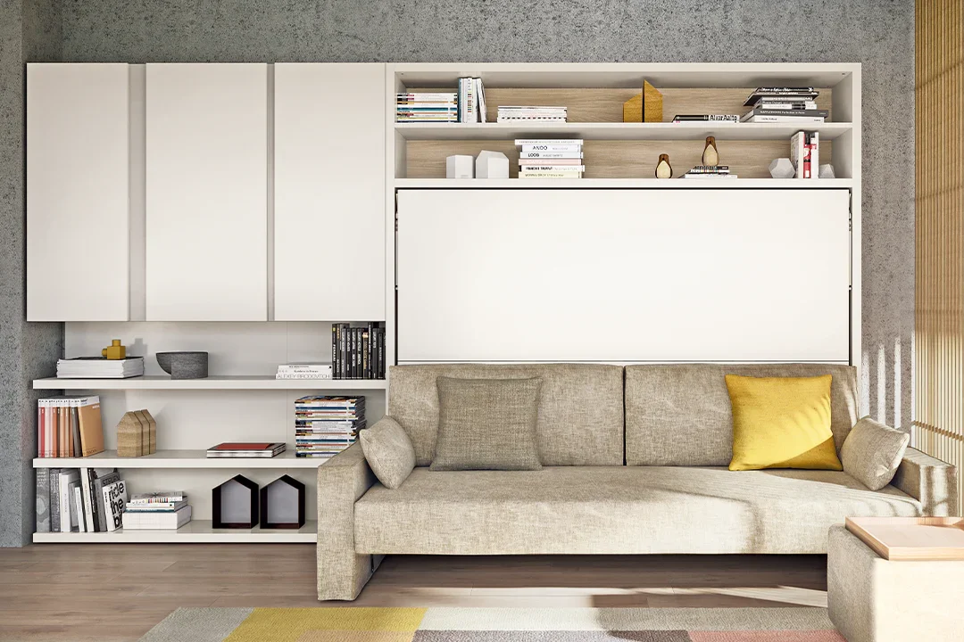 Modern living room with beige sofa, yellow accent pillow, white built-in bookshelf with decorative items and books, concrete wall, wooden flooring, and window with blinds