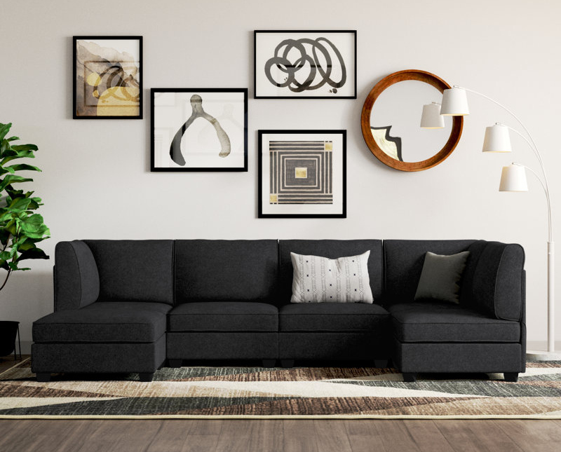 Living room with a black sectional sofa, white and gray pillows, a wall with black picture frames with abstract art, a round wooden mirror, a white multi-arm floor lamp, a potted plant, and a patterned area rug.