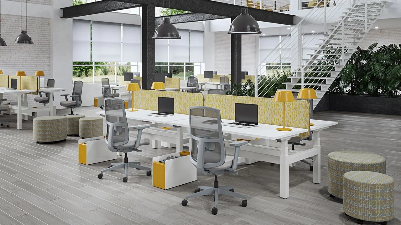 Modern open office space with white desks, gray office chairs, yellow desk lamps, patterned ottomans, and large windows with roller shades. There is a staircase with white railings and large green plants in the background.