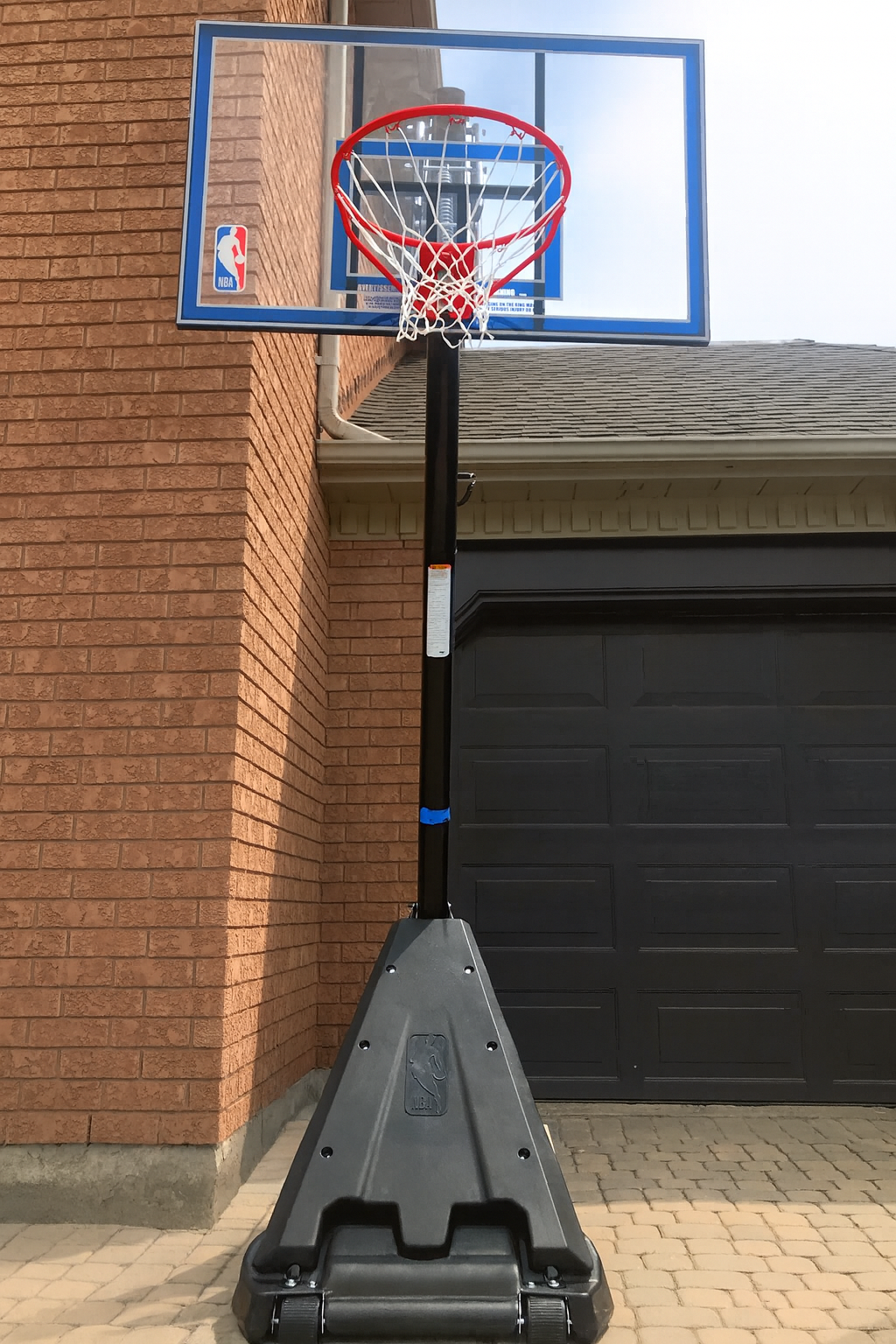 Portable basketball hoop with a blue backboard, red rim, and white net, positioned on a paved driveway in front of a brick house.