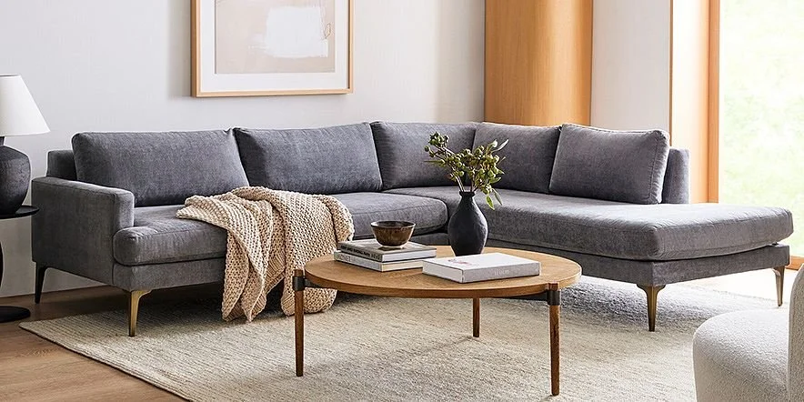Living room with a gray sectional sofa, a wooden coffee table with books and a black vase with greenery, beige throw blanket, cream area rug, window with wooden blinds, and a framed abstract art piece on the wall.