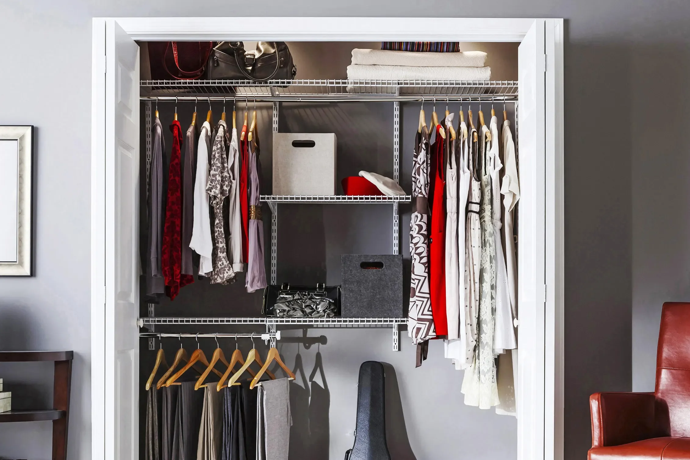 Open closet with various hanging clothes, shelves with folded items and storage boxes, a guitar case leaning against the wall, and part of a red chair visible on the right.