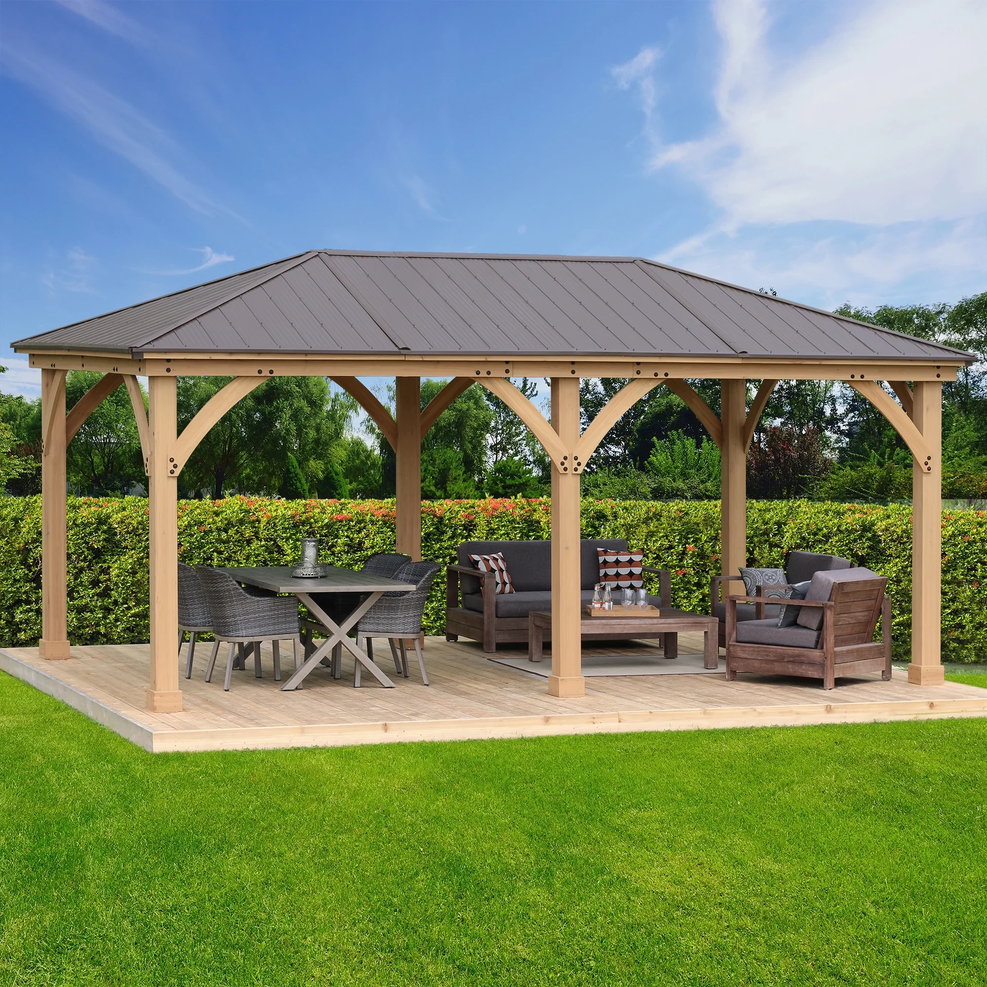 A wooden pavilion with a metal roof located in a grassy area, furnished with outdoor tables and chairs, with trees and shrubbery in the background.
