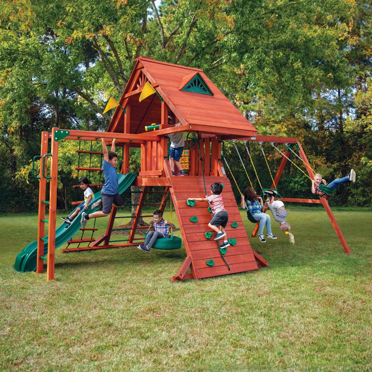 Children playing on a large wooden playground structure with slides, swings, a climbing wall, and a playhouse in a grassy yard surrounded by trees.