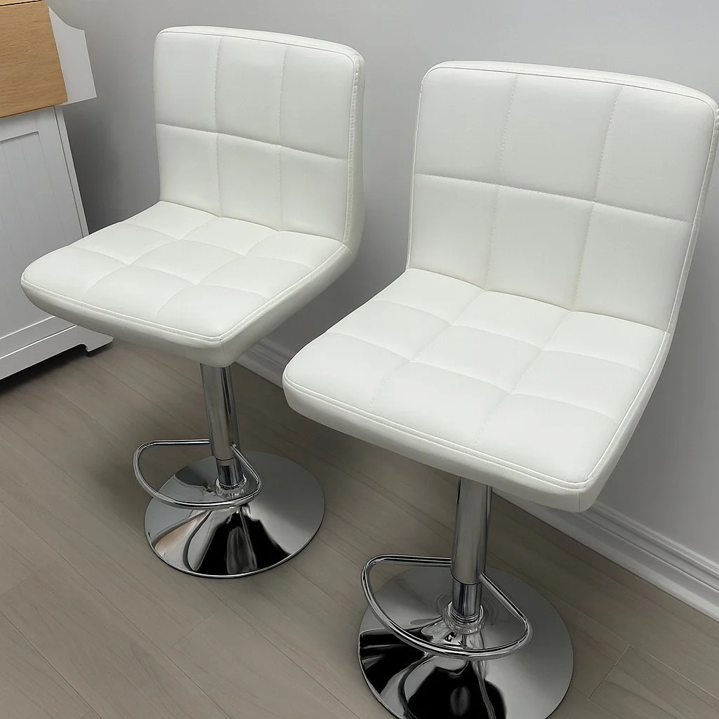 Two white bar stools with cushioned seats and backs, with chrome bases and footrests, positioned on a light wood floor against a plain gray wall.