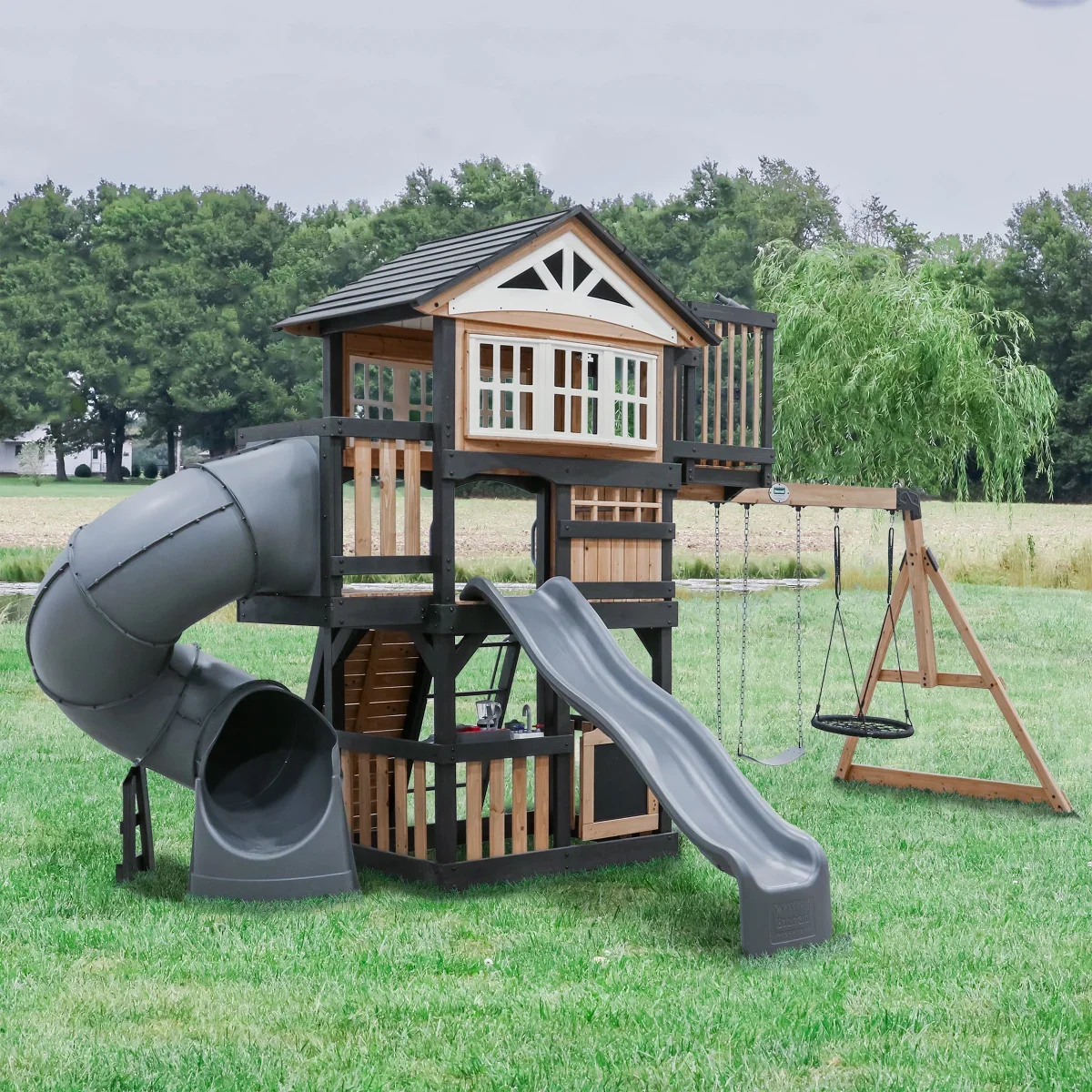 Children's outdoor playset with a treehouse, slide, and swings on a grassy field with trees in the background.