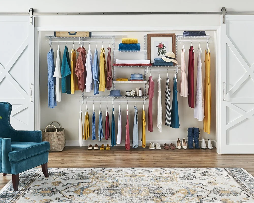 A neatly organized closet with dresses, shirts, and accessories on hanging rods and shelves, a plush teal chair, a woven basket, and a patterned rug on wooden floor.