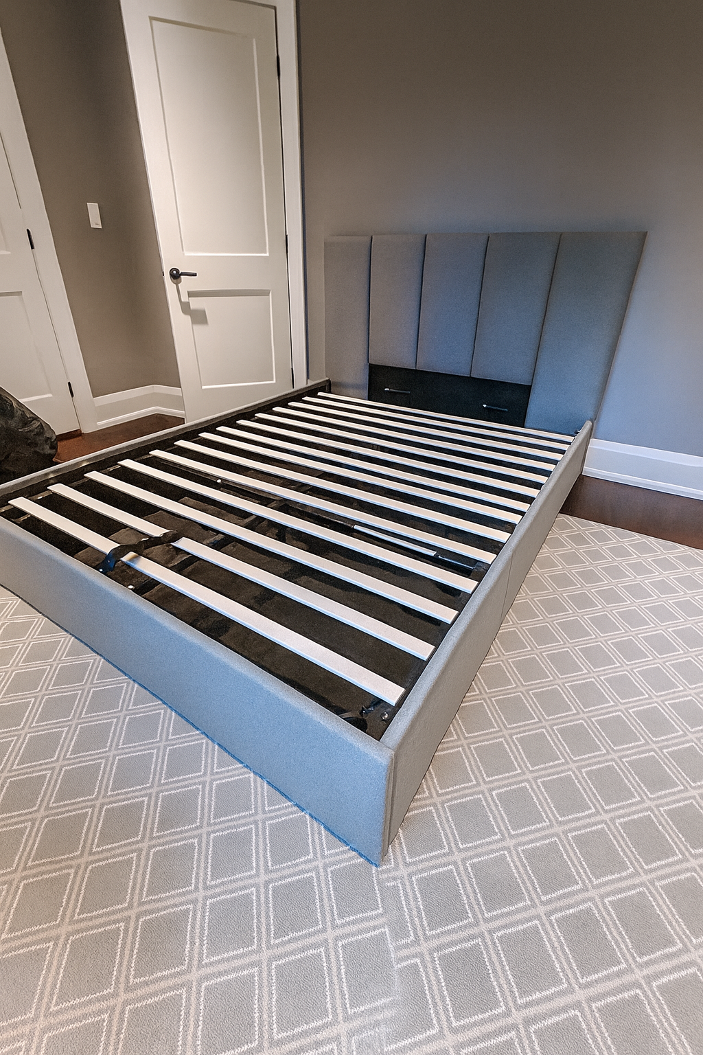 Unassembled bed frame in a bedroom with a patterned carpet, a closed white door, and a headboard leaning against the wall.