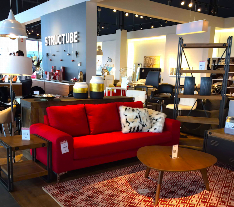 A red sofa with a black and white fur pillow in a furniture store, surrounded by various furniture pieces and decor items, with a sign reading 'STRUCTURE' on the wall in the background.