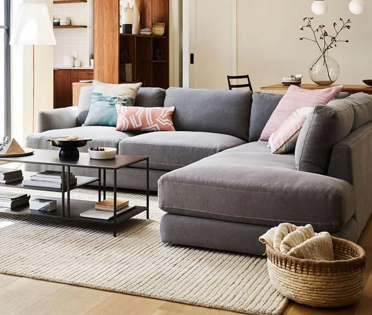 Living room with a light gray sectional sofa decorated with pink and light blue throw pillows, a coffee table with books and decorative items, a beige striped rug, a woven basket with a blanket, a kitchen area, and a dining table with a large glass vase and minimalist decor.