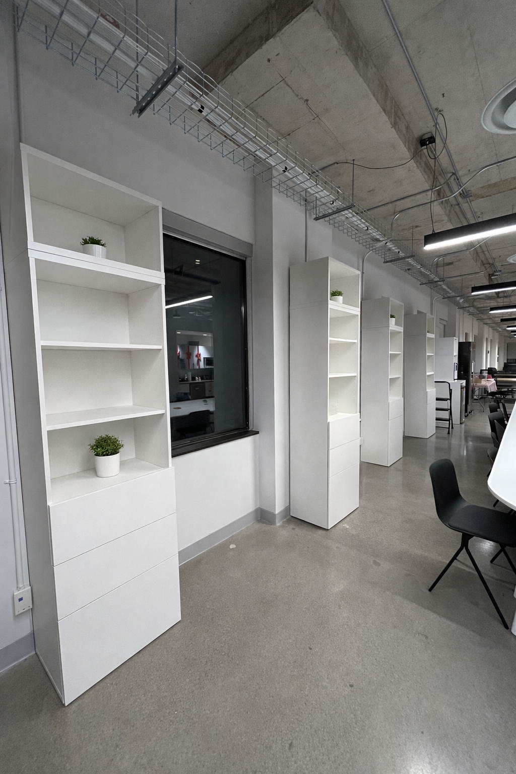 Office space with white shelves, small potted plants, and black chairs under an exposed concrete ceiling.