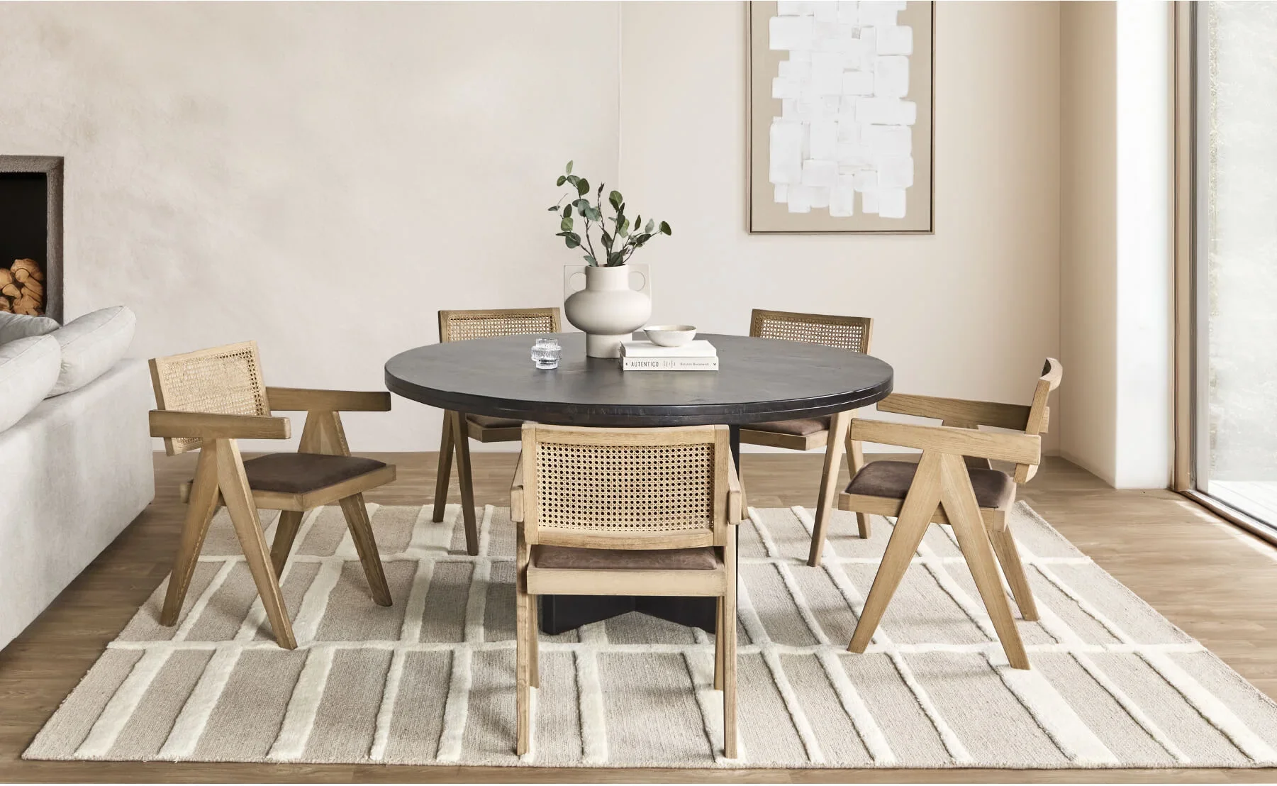 Dining room with a black oval table surrounded by six wooden chairs with woven backs and brown cushions, a white vase with green leaves on the table, a white book, a small bowl, a glass of water, a beige and white striped rug, and a window with sunlight.
