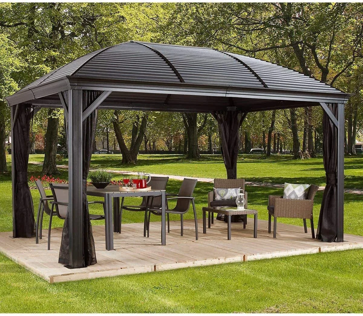 A black metal gazebo with open sides and a curved roof, set on a wooden platform in a park with green grass and trees, decorated with outdoor furniture including a table, chairs, and cushioned armchairs.