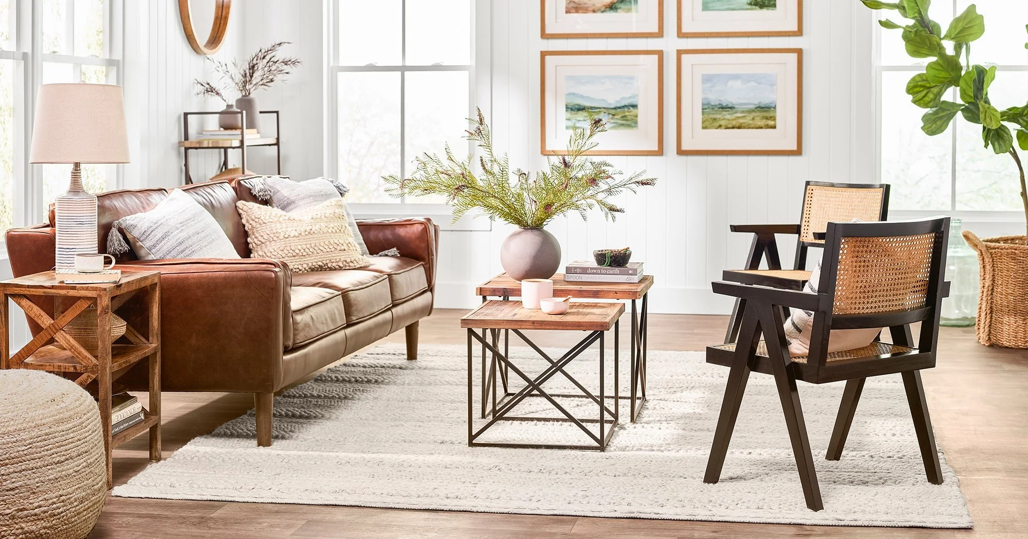A cozy living room with a brown leather sofa, black wooden chairs with woven cane backs, a white area rug, and a central wooden coffee table adorned with a large vase of greenery, candles, and books. The room has white paneled walls, framed landscape paintings, a floor lamp, a small side table, and large windows letting in natural light.