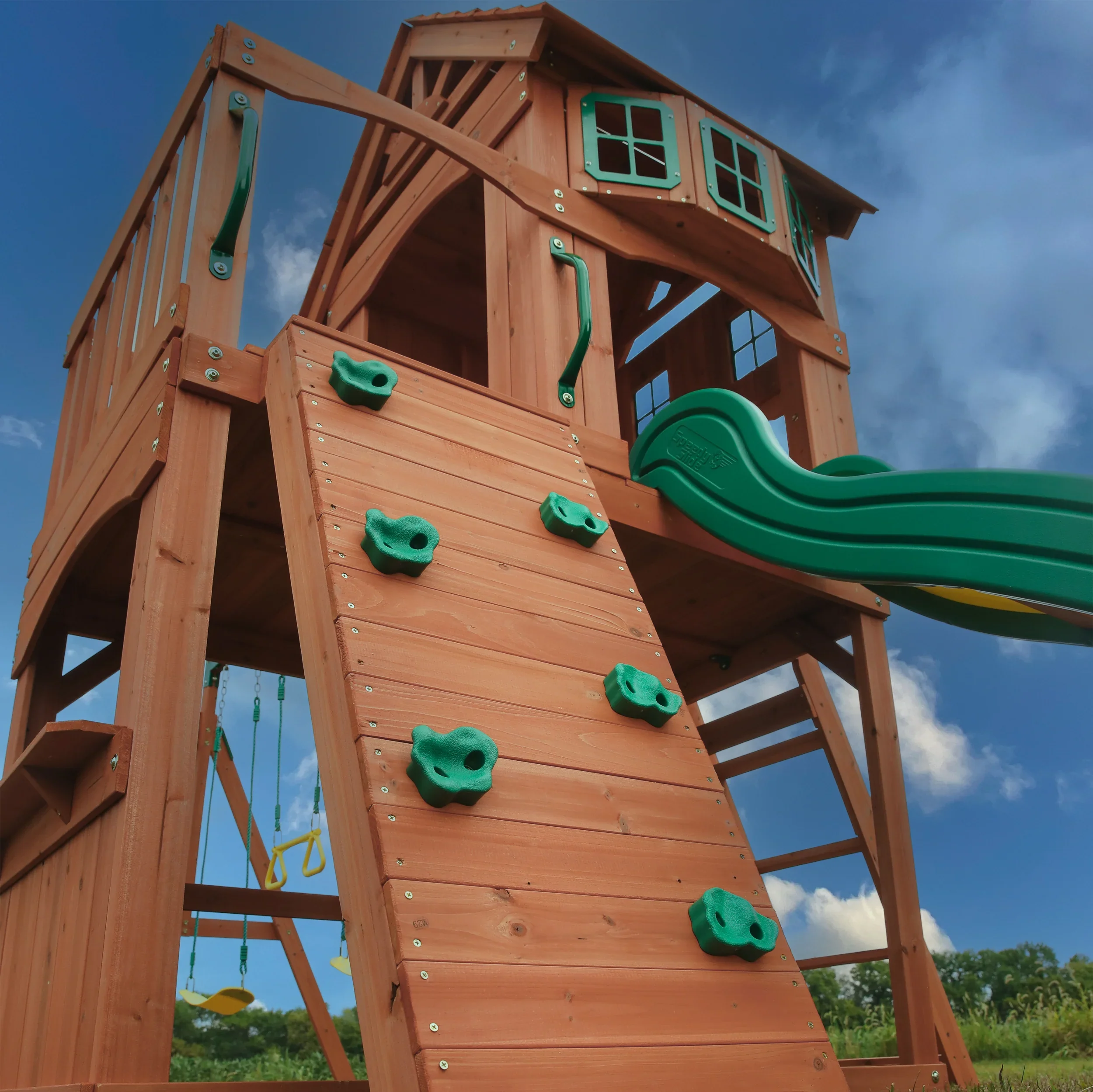 A large wooden playground structure with a green slide, climbing wall, swings, and a small house at the top, set against a blue sky with some clouds.