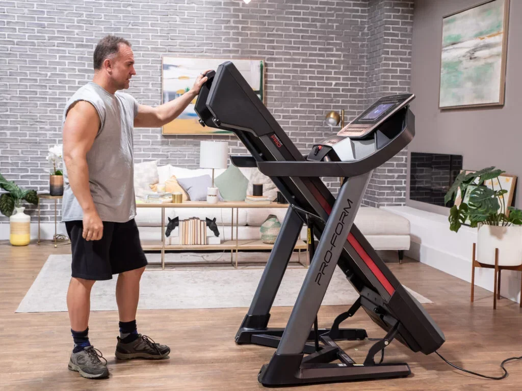A man standing on a treadmill, touching the control panel, in a living room with brick walls, indoor plants, a sofa, and wall art.